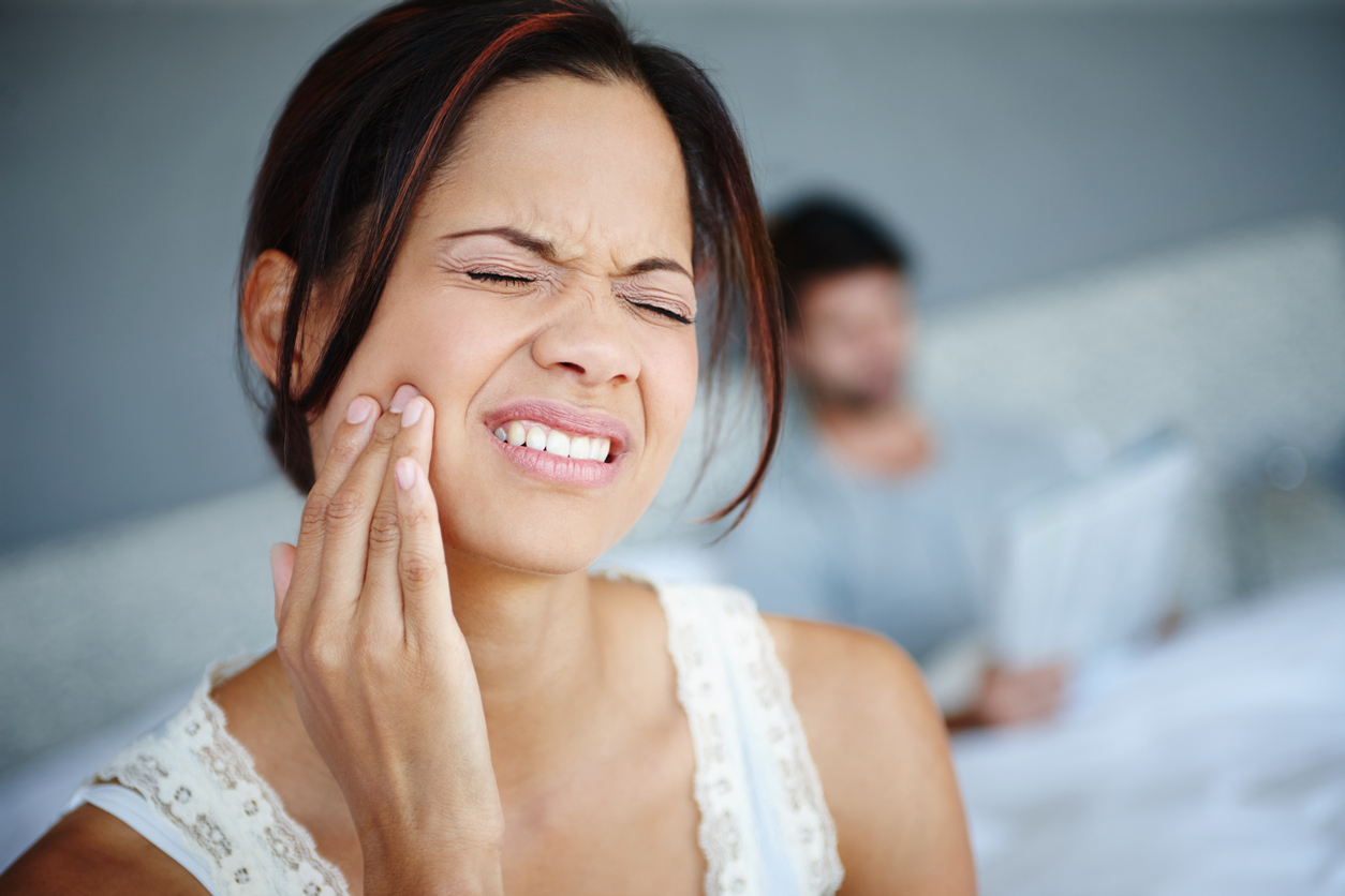 Shot of a woman sitting on the side of her bed with bad toothache with her boyfriend in the background